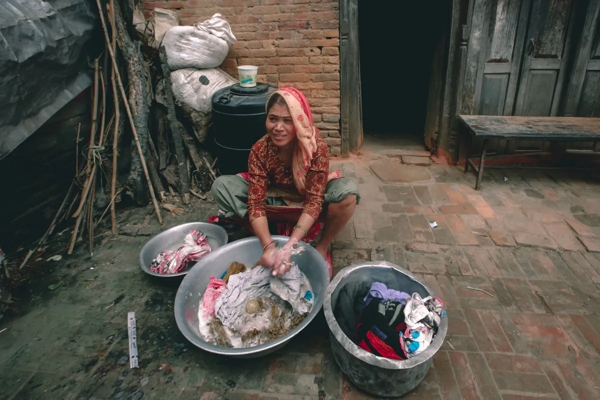 A woman washing clothes by hand in metal basins outside an old stone house