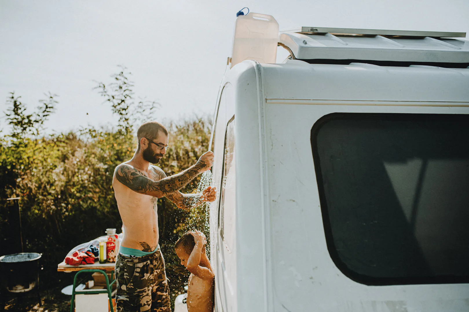 A father giving his young son an outdoor shower beside their campervan, hot water supplied off the grid
