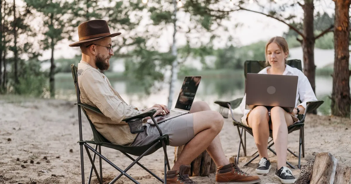 Two people working on laptops in camp chairs at a lakeside campsite