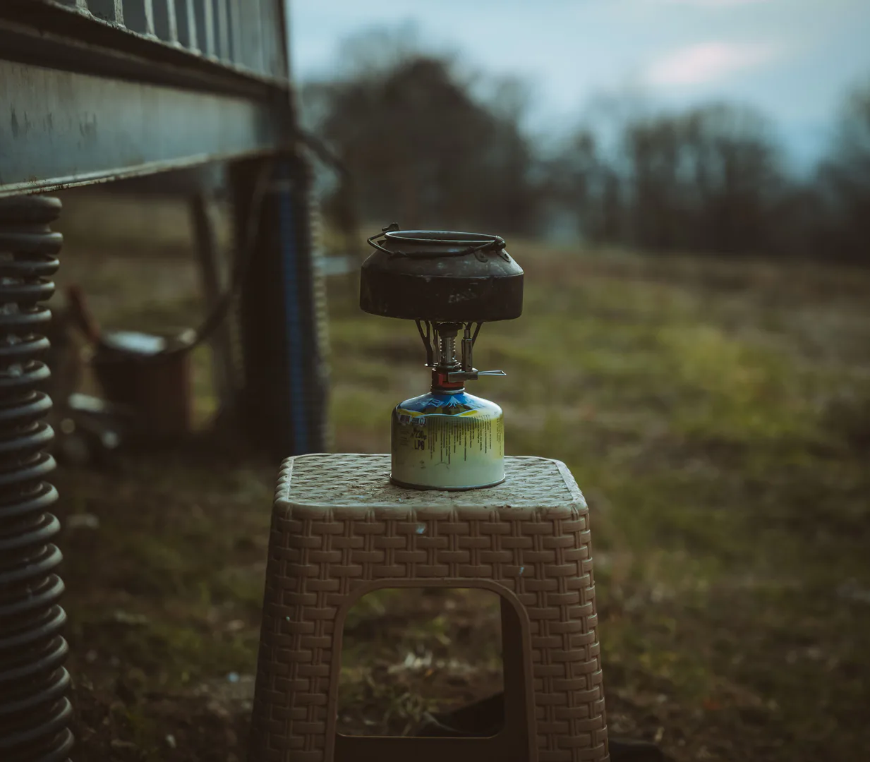 A camping gas burner and cookpot set up on a stool in a rural field