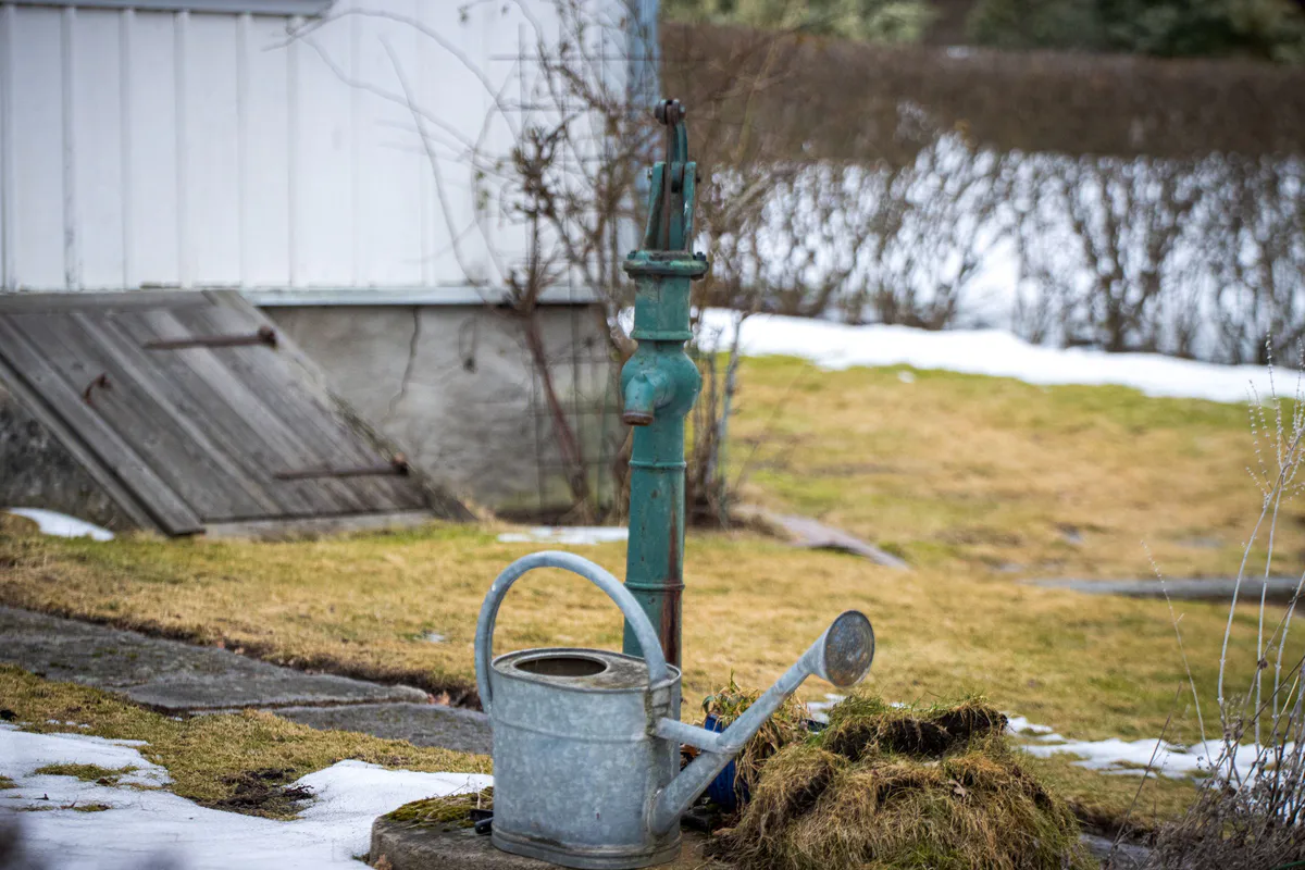 A vintage cast iron garden water pump dusted with snow beside a watering can in a winter garden